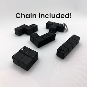 Puede incluir: Cinco piezas de rompecabezas de plástico negro en forma de bloques de Tetris. Las piezas están conectadas entre sí con una cadena de metal y un anillo plateado. "¡Cadena incluida!" está escrito en texto blanco sobre un fondo blanco.