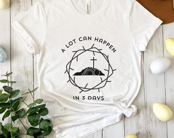 Pode incluir: Camiseta branca com um gráfico preto que diz "Muita coisa pode acontecer em 3 dias". O gráfico apresenta uma coroa de espinhos, uma cruz e um túmulo.