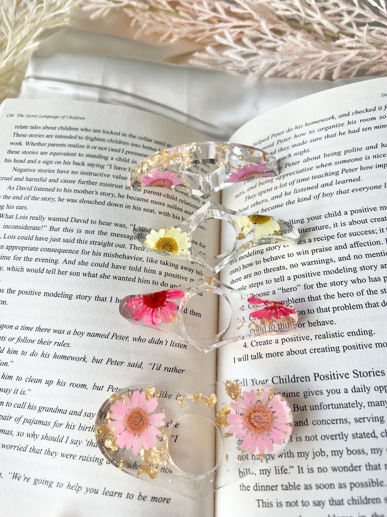 K&ouml;nnte beinhalten: Drei Harzringe mit Blattgold und gepressten Blumen. Die Ringe sind in Rosa- und Wei&szlig;t&ouml;nen gehalten. Die Ringe liegen auf einem Buch mit dem sichtbaren Text "The Secret Language of Children".