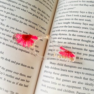 K&ouml;nnte beinhalten: Nahaufnahme eines pinkfarbenen und wei&szlig;en Blumen, die in einem klaren Harz-Lesezeichen gepresst ist. Das Lesezeichen ist in Form einer Brille geformt und hat goldene Akzente. Das Lesezeichen liegt auf einem ge&ouml;ffneten Buch mit dem Text "Ray, do you know how to..." sichtbar.