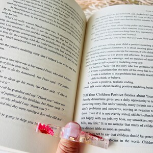 K&ouml;nnte beinhalten: Nahaufnahme eines rosa-goldenen Lesezeichens mit einer gepressten Trockenblume. Das Lesezeichen liegt auf einem ge&ouml;ffneten Buch mit Text &uuml;ber positive Vorbildgeschichten f&uuml;r Kinder.