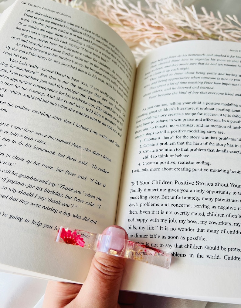 K&ouml;nnte beinhalten: Nahaufnahme eines rosa-goldenen Lesezeichens mit einer gepressten Trockenblume. Das Lesezeichen liegt auf einem ge&ouml;ffneten Buch mit Text &uuml;ber positive Vorbildgeschichten f&uuml;r Kinder.