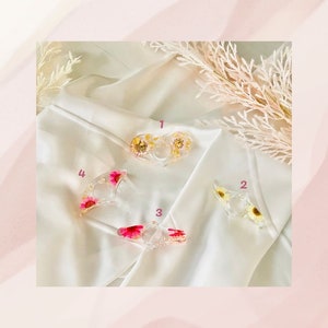 K&ouml;nnte beinhalten: Vier transparente Ringhalter aus Harz mit gepressten Blumen. Die Halter haben die Form eines Rings, eines Halbmondes und zweier Herzen. Die Blumen sind rosa, wei&szlig; und gelb.