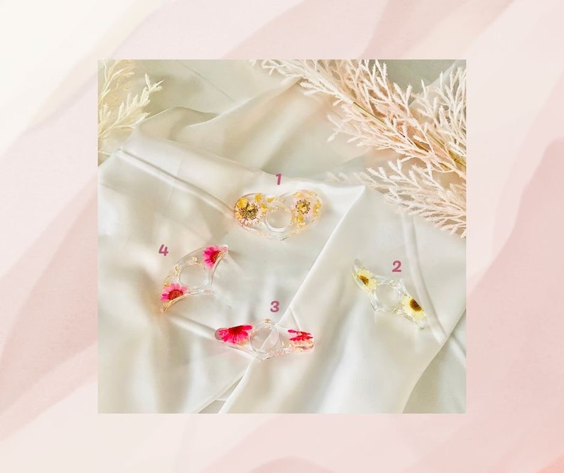 K&ouml;nnte beinhalten: Vier transparente Ringhalter aus Harz mit gepressten Blumen. Die Halter haben die Form eines Rings, eines Halbmondes und zweier Herzen. Die Blumen sind rosa, wei&szlig; und gelb.