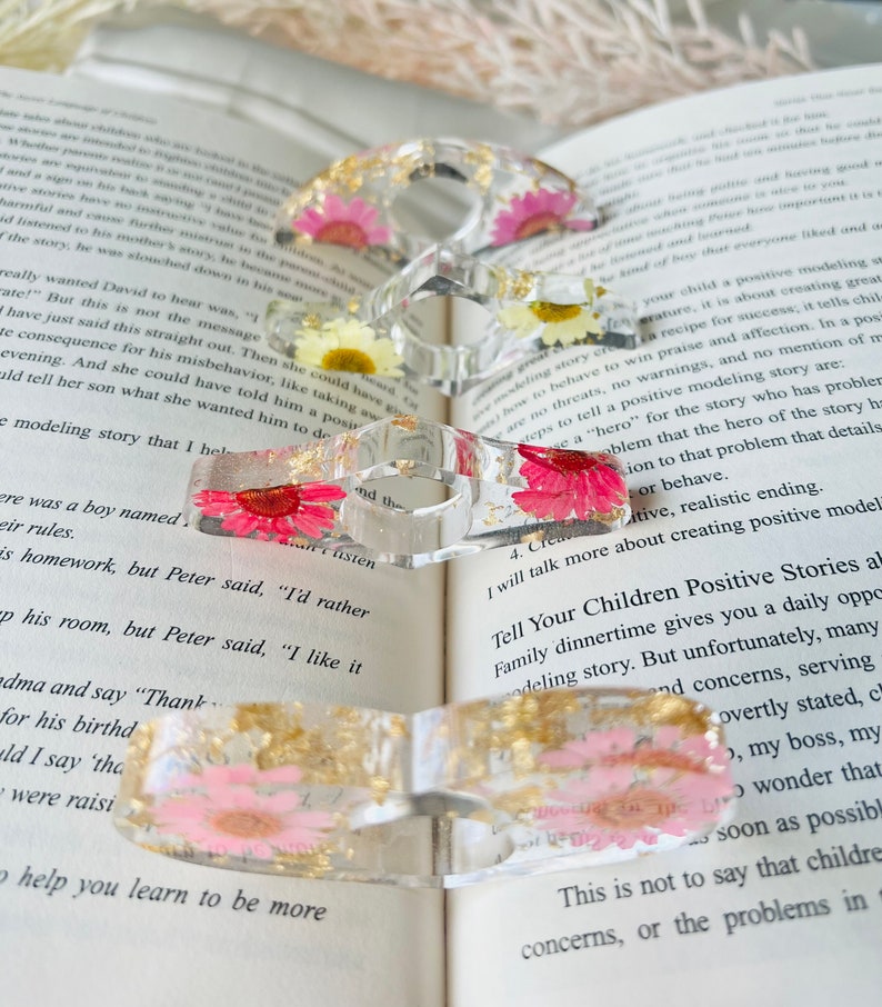 K&ouml;nnte beinhalten: Vier transparente Harzringe mit eingebetteten Trockenblumen und Blattgold. Die Ringe sind &uuml;bereinander gestapelt und liegen auf einem Buch.