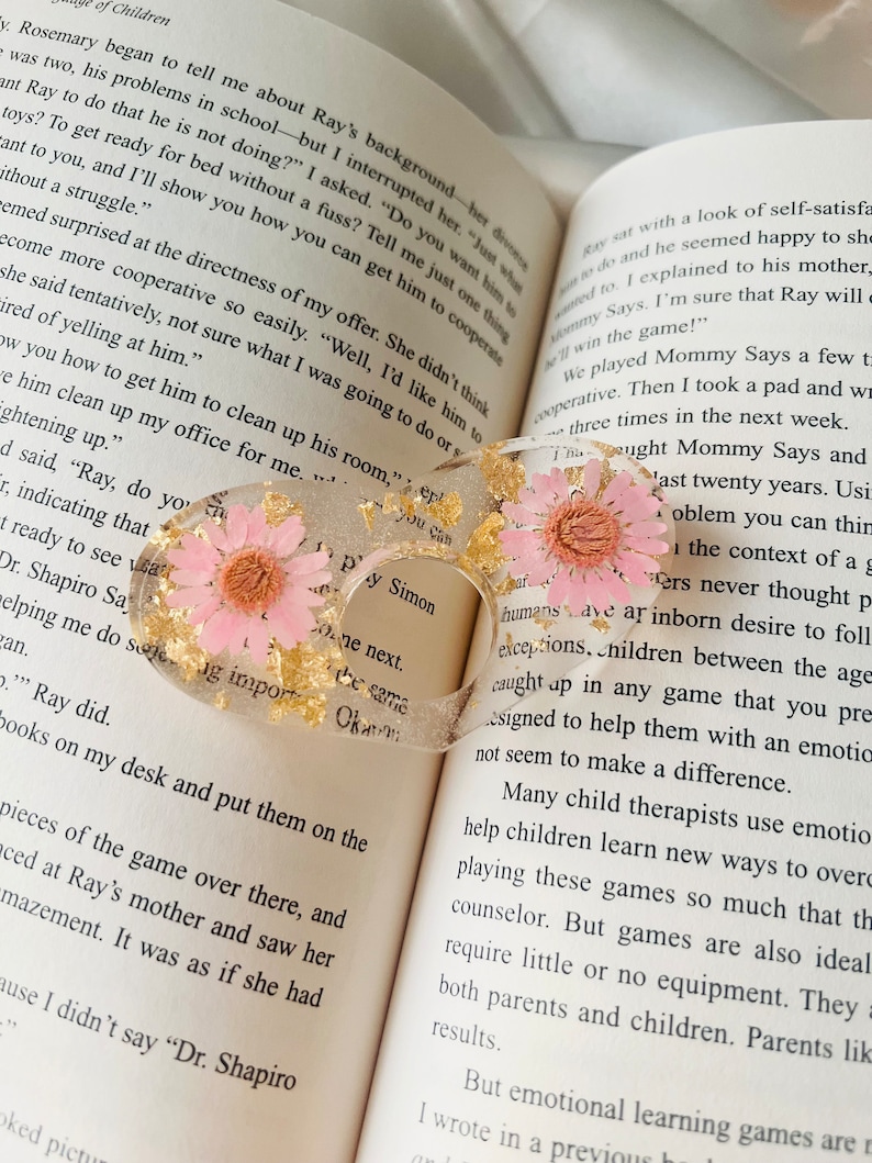 K&ouml;nnte beinhalten: Nahaufnahme eines Buches mit einem rosafarbenen und goldenen Harz-Lesezeichen mit gepressten Blumen. Das Lesezeichen tr&auml;gt den Text "Py Simon" und "You Can". Das Buch ist auf einer Seite &uuml;ber ein Kind namens Ray und seine Mutter ge&ouml;ffnet.