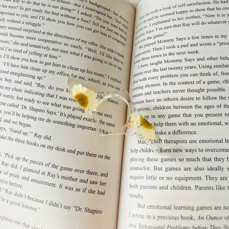 K&ouml;nnte beinhalten: Nahaufnahme eines Buches mit dem Text "Mommy Says" und "Dr. Shapiro Says." Das Buch ist ge&ouml;ffnet und ein Paar klarer herzf&ouml;rmiger Brillen mit getrockneten wei&szlig;en Blumen liegen auf den Seiten.