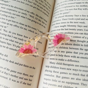 K&ouml;nnte beinhalten: Ein transparentes herzf&ouml;rmiges Lesezeichen mit goldenem Glitzer und gepressten rosa Bl&uuml;ten. Das Lesezeichen liegt auf einem ge&ouml;ffneten Buch mit Text &uuml;ber ein Spiel namens "Mommy Says".