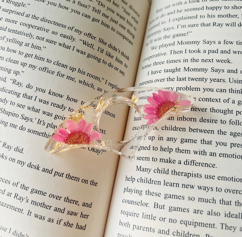 K&ouml;nnte beinhalten: Ein transparentes herzf&ouml;rmiges Lesezeichen mit goldenem Glitzer und gepressten rosa Bl&uuml;ten. Das Lesezeichen liegt auf einem ge&ouml;ffneten Buch mit Text &uuml;ber ein Spiel namens "Mommy Says".
