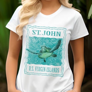 May include: White t-shirt with a turquoise graphic of a sea turtle swimming in the ocean. The graphic is in the style of a vintage postage stamp and says "ST. JOHN U.S. VIRGIN ISLANDS".