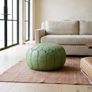 May include: A round, sage green leather pouf featuring a stitched petal-like pattern on its top surface. The pouf is positioned on a textured, rust-colored area rug in a room with light-colored walls, a beige sofa, and a brown leather chair.