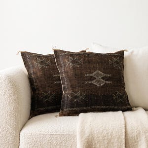 May include: Two square scatter cushions featuring a dark brown, textured fabric with white embroidered geometric diamond patterns and frayed edges, positioned alongside a plain, solid off-white scatter cushion on a textured cream-coloured sofa.