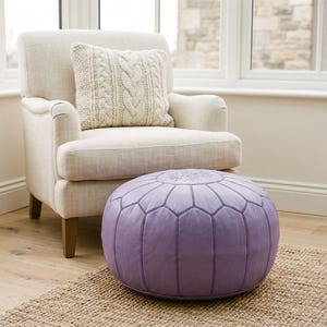 May include: A round, lavender-colored leather pouf featuring stitched paneling and a central floral embroidery design, positioned in front of an off-white upholstered armchair with a matching cream-colored cable knit throw pillow.