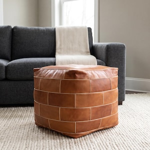 May include: A square, tan leather ottoman featuring a brick-like pattern of rectangular panels stitched together with light-colored thread. The piece has a structured, boxy shape with visible seams defining each individual leather section.