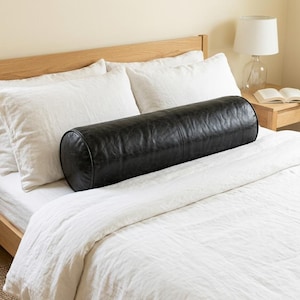 May include: A black cylindrical bolster pillow made of a smooth, leather-like material. The pillow features a horizontal seam running across its center and is positioned horizontally across the middle of a bed with white bedding and pillows.