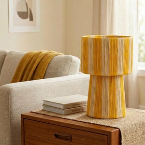 May include: A decorative table lamp with a cylindrical shade and base, featuring vertical stripes of yellow and cream-colored yarn. The lamp sits on a wooden nightstand with two books. A mustard-colored throw blanket is draped over a light gray sofa.