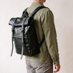 May include: A black leather roll-top backpack featuring two vertical straps with gold-toned metal buckles, a top handle, and a horizontal zippered front pocket. The bag has adjustable shoulder straps and a smooth, matte finish on the exterior surface.