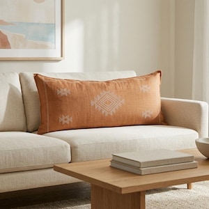 May include: A rectangular lumbar pillow in a burnt orange hue featuring a centered white geometric diamond pattern and smaller matching diamond motifs on the sides, crafted with a textured fabric finish and placed on a light beige sofa.