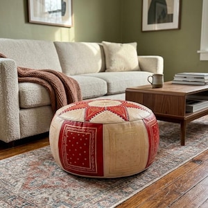 May include: A round, patterned ottoman with red and cream-coloured panels sits on a patterned rug in a living room. The ottoman has a starburst design on top. A beige sofa and wooden coffee table are also in the room.