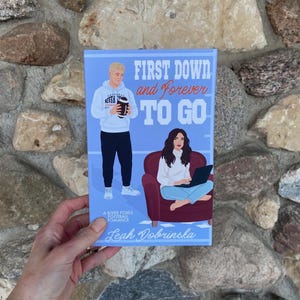 May include: A book cover with the title "FIRST DOWN and Forever TO GO" featuring illustrations of a football player and a woman with a laptop. The author's name, Leah Dobrinska, is at the bottom. The book is a River Foxes Football Romance.