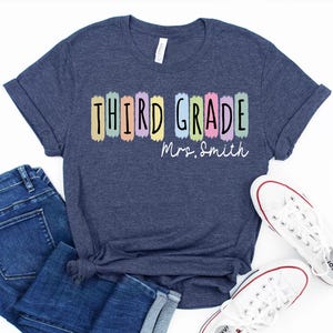 May include: A heather navy blue t-shirt featuring the text "THIRD GRADE" in black letters over colorful pastel brushstroke rectangles, with the white cursive text "Mrs. Smith" underneath. The shirt is paired with blue denim jeans and white sneakers.