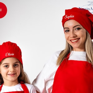 May include: Two red chef hats with the names "Sophia" and "Olivia" embroidered on them. The hats are worn by a woman and a girl, both wearing white shirts and red aprons.
