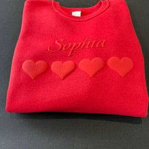 May include: A red sweatshirt with the name "Sophia" embroidered in a matching red thread. Four red hearts are aligned horizontally below the name. The sweatshirt is folded neatly on a dark surface.