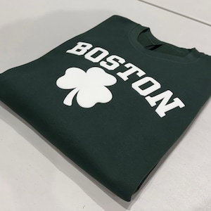 May include: A dark green crewneck sweatshirt featuring the word BOSTON printed in large white capital letters above a white three-leaf clover graphic. The garment is folded neatly, showcasing the screen-printed design on the front chest area.