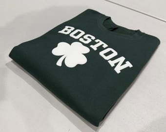 May include: A dark green crewneck sweatshirt featuring the word BOSTON printed in large white capital letters above a white three-leaf clover graphic. The garment is folded neatly, showcasing the screen-printed design on the front chest area.