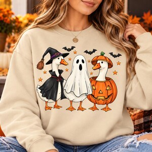 May include: A beige crewneck sweatshirt featuring a graphic print of three white geese dressed for Halloween. One goose wears a black witch hat and cape, the middle goose is covered by a white ghost sheet, and the third goose sits inside a carved pumpkin.