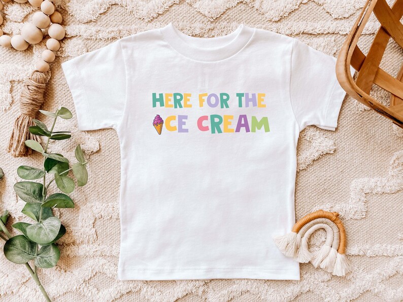 Pu&ograve; includere: Maglietta bianca con una grafica colorata che dice "Here for the Ice Cream" con un cono gelato disegnato.