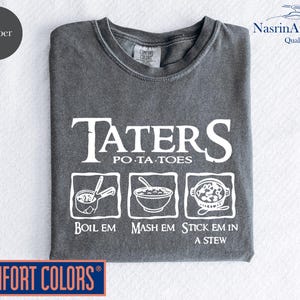 May include: A gray t-shirt with the text "TATERS POTATOES" in white. Below the text are three illustrations of different ways to prepare potatoes: "BOIL EM", "MASH EM", and "STICK EM IN A STEW". The shirt is from Comfort Colors.