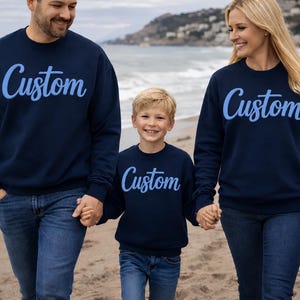 May include: Three people wearing matching navy blue sweatshirts with the word "Custom" in light blue script. They are walking on a beach, holding hands. The sweatshirts are long-sleeved.