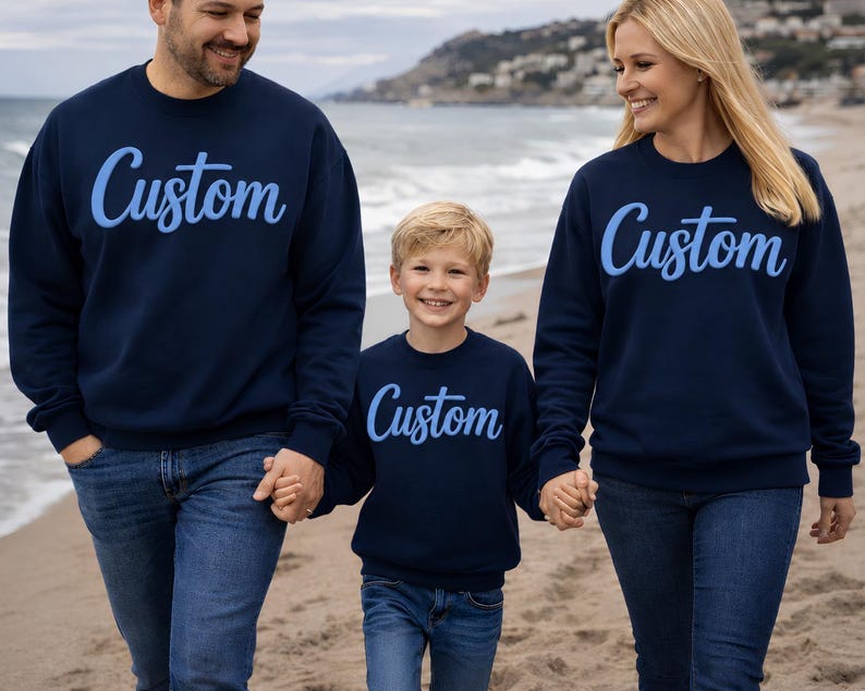 May include: Three people wearing matching navy blue sweatshirts with the word "Custom" in light blue script. They are walking on a beach, holding hands. The sweatshirts are long-sleeved.