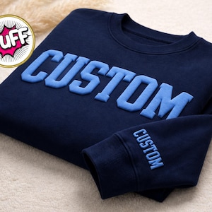 May include: Navy blue sweatshirt with the word "CUSTOM" in light blue, raised lettering across the chest. The sleeve cuff also features the word "CUSTOM" in light blue. The sweatshirt is folded on a light-colored surface.