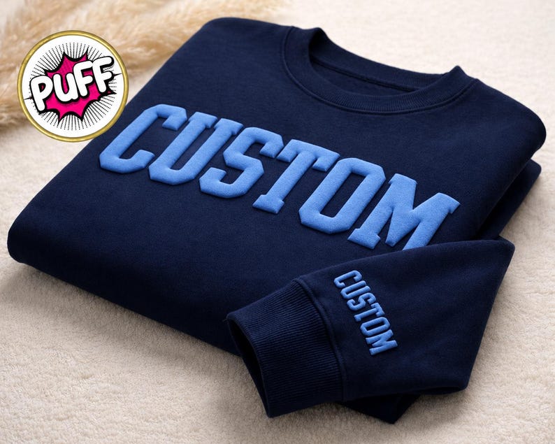 May include: Navy blue sweatshirt with the word "CUSTOM" in light blue, raised lettering across the chest. The sleeve cuff also features the word "CUSTOM" in light blue. The sweatshirt is folded on a light-colored surface.