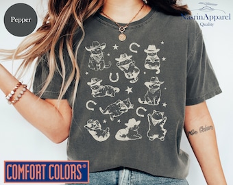 Può includere: Una maglietta grigia scura con una stampa grafica di gatti che indossano cappelli da cowboy e bandane. La grafica include anche stelle e ferri di cavallo. La maglietta ha il testo "Comfort Colors" nell'angolo in basso a sinistra e "NasrinApparel Quality" nell'angolo in alto a destra.