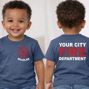 May include: A heather navy short-sleeve t-shirt featuring a red fire department emblem and the name NICOLAS on the front left chest. The back displays the white text YOUR CITY and DEPARTMENT with the red text FIRE centered in between.