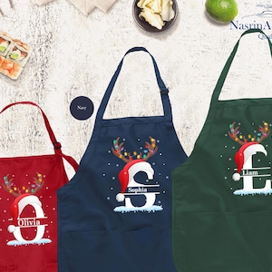 May include: Three personalized Christmas aprons in red, navy blue, and kelly green. Each apron features a reindeer with a Santa hat and a personalized letter with a name. The aprons are perfect for holiday baking and cooking.