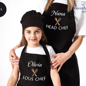 May include: Two black aprons with white text. The larger apron says "Nana Head Chef" with crossed wooden spoons and a whisk. The smaller apron says "Olivia Sous Chef" with crossed wooden spoons and a whisk.