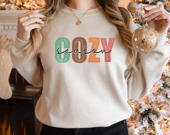 May include: A white sweatshirt with the text "Cozy Season" in colorful, retro-style lettering. The letters are outlined in white and have a distressed, vintage look.