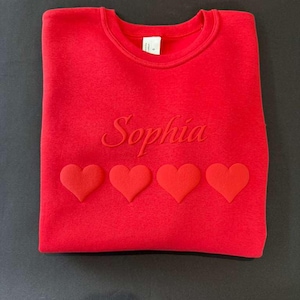May include: A red sweatshirt with the name "Sophia" embroidered in a matching red thread. Below the name are four raised heart-shaped appliqu&eacute;s. The sweatshirt is folded neatly on a dark surface.