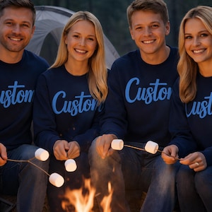 May include: Four people wearing navy blue sweatshirts with the word "Custom" in light blue script. They are gathered around a campfire, roasting marshmallows on sticks. The scene is set outdoors, with a tent visible in the background.