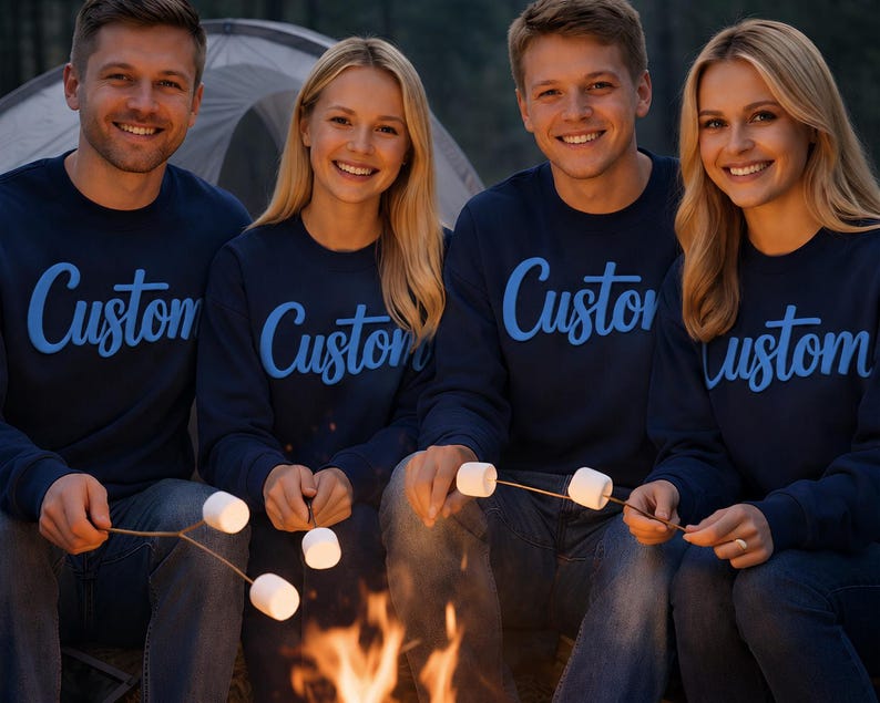 May include: Four people wearing navy blue sweatshirts with the word "Custom" in light blue script. They are gathered around a campfire, roasting marshmallows on sticks. The scene is set outdoors, with a tent visible in the background.