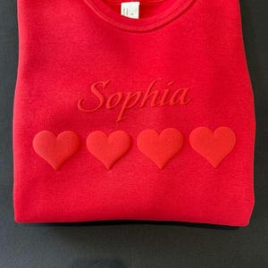 May include: A red sweatshirt with the name "Sophia" embroidered across the chest. Four red hearts are aligned below the name. The sweatshirt is folded and displayed on a black surface.