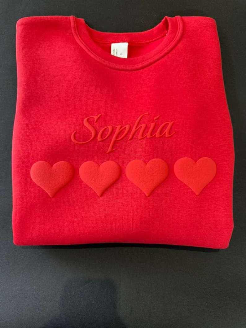 May include: A red sweatshirt with the name "Sophia" embroidered across the chest. Four red hearts are aligned below the name. The sweatshirt is folded and displayed on a black surface.