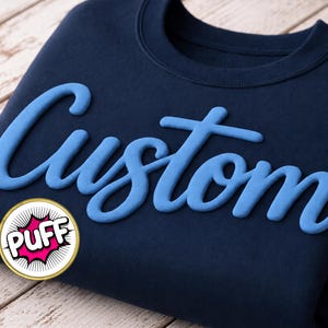 May include: Navy blue sweatshirt with the word "Custom" in light blue, stylized script. A circular graphic with the word "PUFF" in a comic book style is at the bottom left. The sweatshirt is folded on a light wood surface.