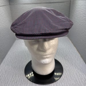 May include: A flat cap in a burgundy, teal, and grey striped pattern. The cap is displayed on a mannequin head, highlighting its shape and design. It features a rounded crown and a small, stiff brim.