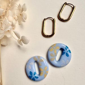 Peut inclure: Une collection de boucles d'oreilles. Deux paires de boucles d'oreilles sont présentées. Une paire est constituée de créoles ovales dorées. L'autre paire est composée de boucles d'oreilles circulaires bleu clair avec une ouverture ovale centrale, décorées de motifs floraux dorés et bleus.