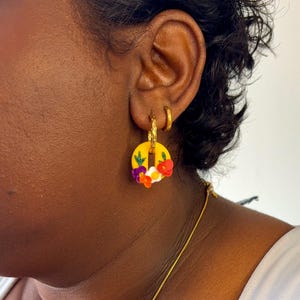 Peut inclure: Une boucle d'oreille créole dorée avec une breloque circulaire jaune ornée de petites fleurs colorées en rouge, blanc, violet et vert. La boucle est portée au lobe, accompagnée d'un anneau doré simple sur le cartilage supérieur.